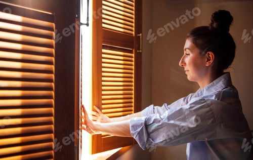 Preview: A young woman opens a wooden blind and looks out the window