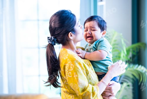 Preview: Indian Mom gently holding sleeping infant boy on shoulder, bonding quietly in bright modern home