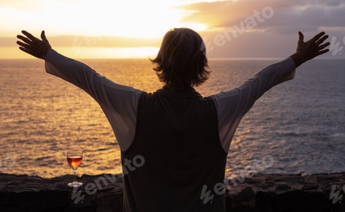 Preview: Rear view of senior woman's silhouette standing in front of the sea with outstretched arms at sunset