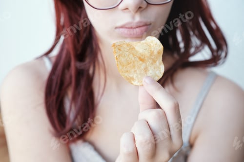 Preview: Red-haired girl holds one potato chip in her hand near her mouth, close-up.