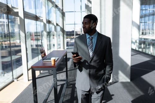 Preview: Thoughtful young businessman with mobile phone looking away standing in office