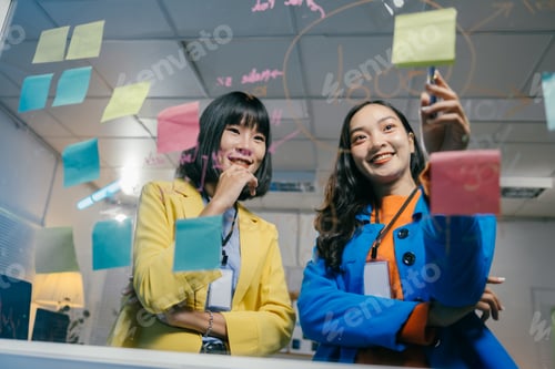 Preview: Two asian businesswomen are using a glass wall with sticky notes and marker pens to brainstorm new i