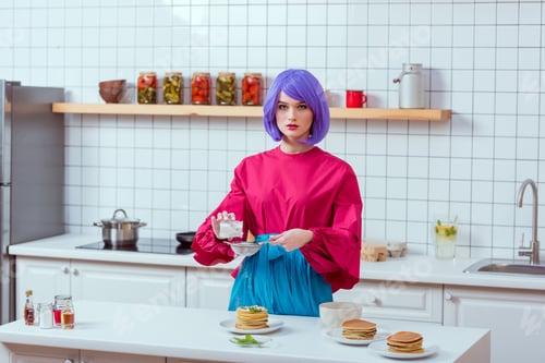 Preview: housewife with purple hair and colorful clothes looking at camera while preparing pancakes in