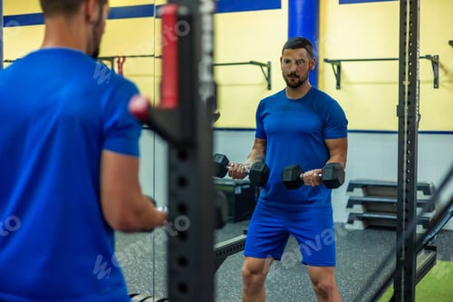 Preview: young fitness athlete doing bicep exercises with dumbbells while reflecting in gym mirror