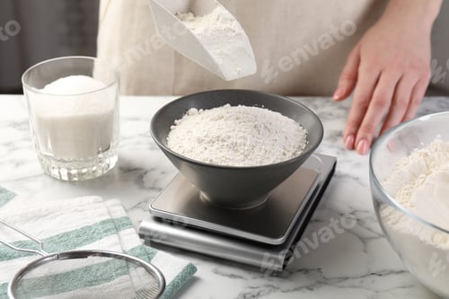 Preview: Woman putting sugar into bowl on digital kitchen scale at white marble table, closeup