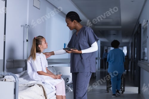 Preview: Diverse female doctor talking with girl using tablet in hospital corridor