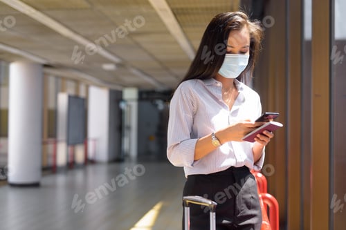 Preview: Young Asian tourist woman with mask using phone and holding passport at the airport