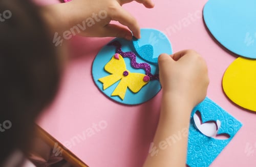 Preview: The hands of a little caucasian girl stick a hat sticker on a blue felt egg with a lilac zigzag