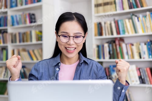 Preview: Excited woman celebrating success while working on laptop in library setting