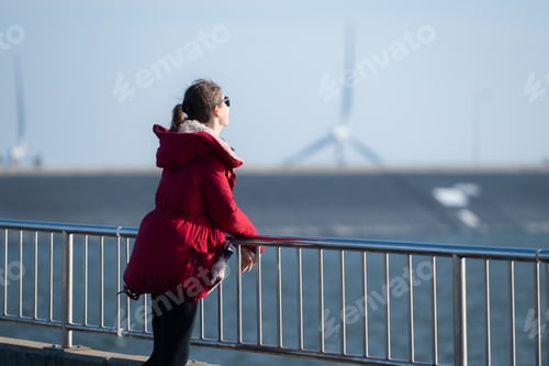 Preview: Woman in Red Jacket by Water on Sunny Day