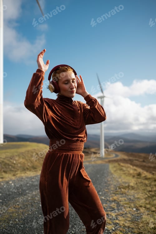 Preview: Young sporty woman listening music with headphones and enjoying nature near wind turbines