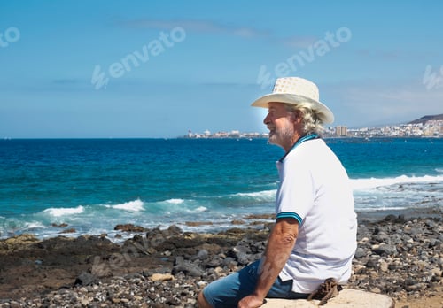 Preview: Smiling senior man on the pebble beach looking at the horizon over water. Blue sky and sea