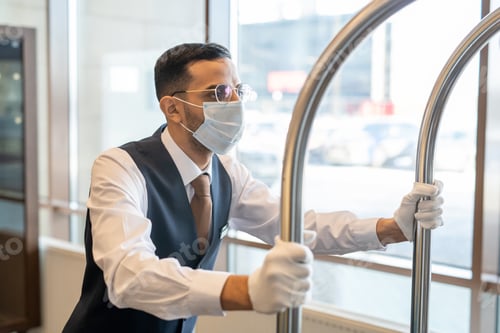 Preview: Young porter in uniform, eyeglasses and protective mask pushing cart with baggage along corridor