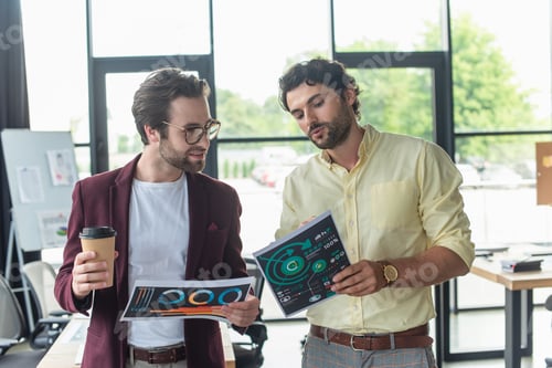 Preview: Businessman holding paper and talking to smiling colleague with coffee to go in office