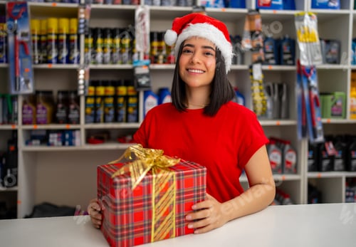 Preview: Smiling woman wearing santa hat offering a christmas gift in a retail store