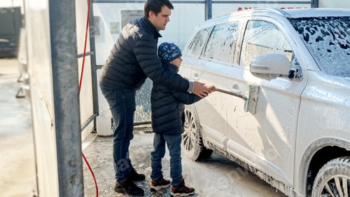 Preview: Young smiling man with son washing car outdoors with brush and foam. Automobile care, dirty car.