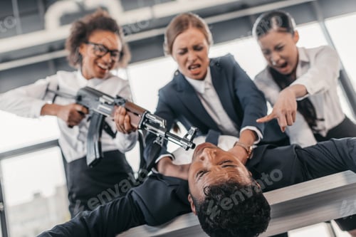 Preview: Three young angry businesswomen with rifle punishing businessman lying on table, business team