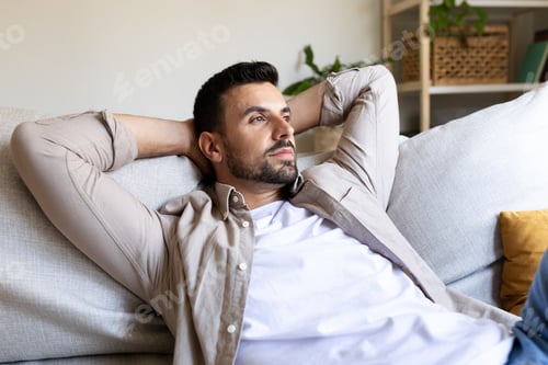 Preview: Pensive Caucasian man relaxing on the sofa with hands behind the head looking away.