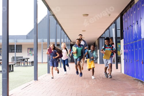 Preview: Diverse male teacher with happy children running in elementary school corridor, copy space