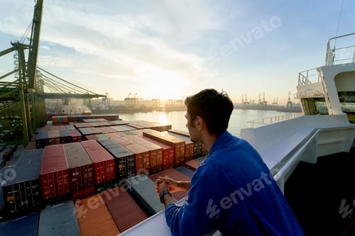 Preview: A person stands on a ship, gazing out at a harbor filled with shipping containers during sunrise.