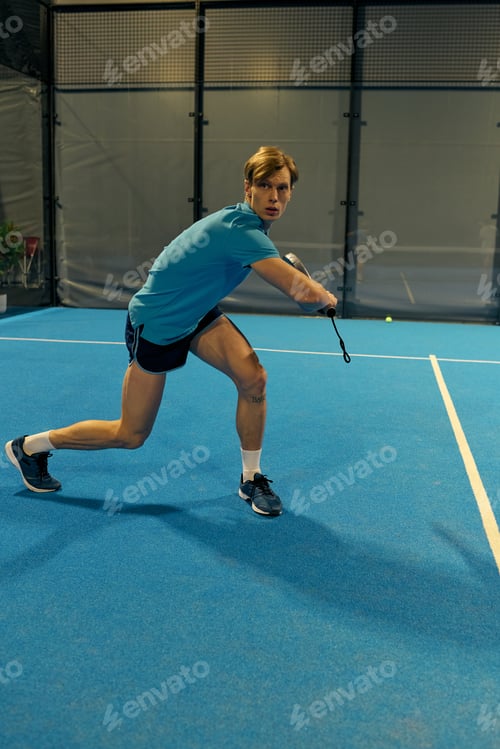 Preview: Young sportsman skillfully playing padel on a vibrant blue court during daytime