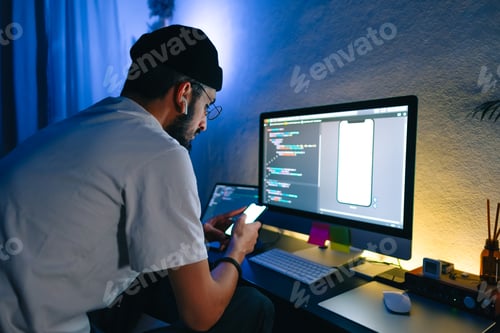 Preview: Young man mobile developer writes program code on a computer and check app in smartphone