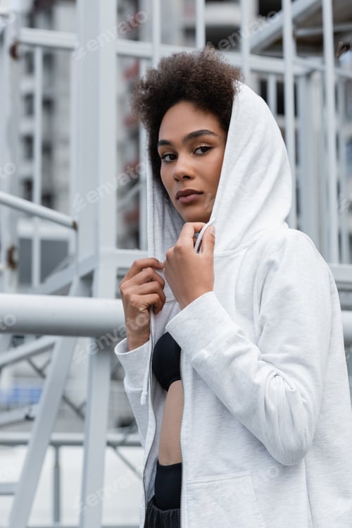 Preview: brunette african american woman in white hoodie looking at camera on urban stadium