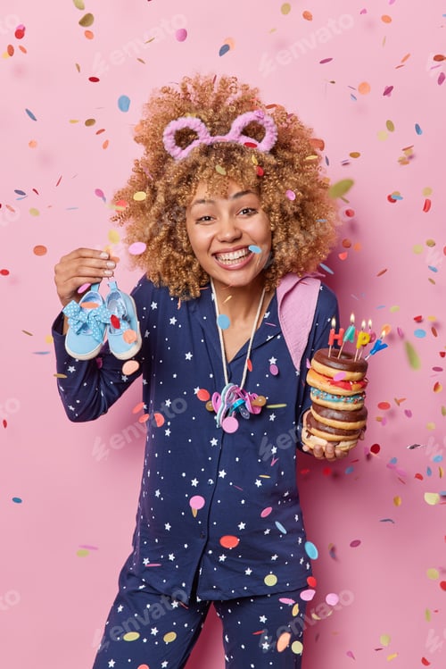 Preview: Happy pregnancy concept. Positive curly haired woman celebrates birthday at home holds shoes and pil