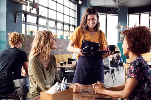 Preview: Two Female Friends Sitting At Table In Coffee Shop Being Served By Waitress