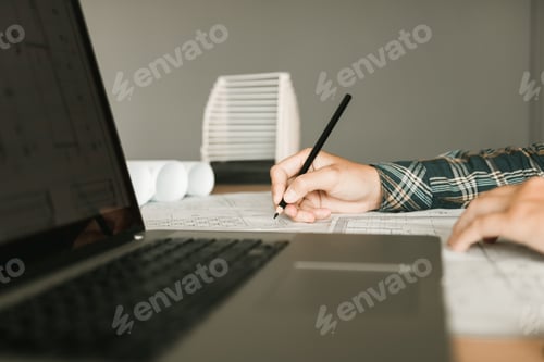 Preview: Architect working on blueprint in workplace with laptop and drawing.