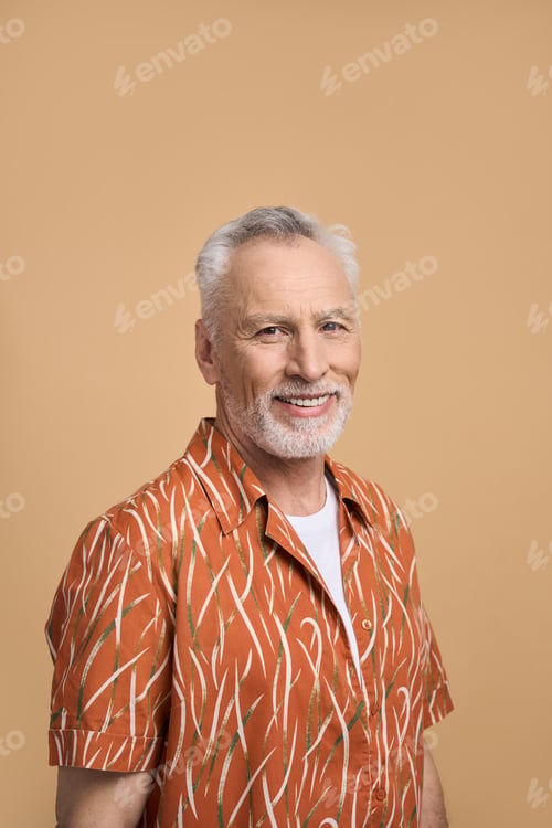 Preview: Handsome cheerful bearded senior man in bright orange shirt, smiling at camera, isolated on beige