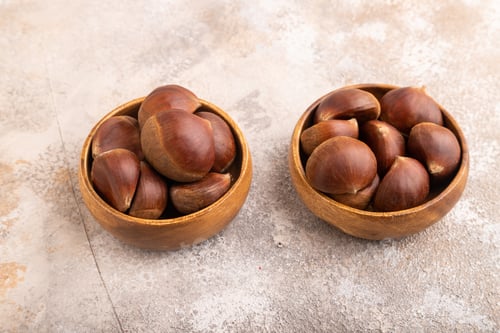 Preview: wooden bowl with raw edible Сhestnuts on brown concrete, side view, close up, minimalism