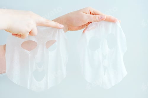 Preview: Female hands holding two moisturizing mask sheets on gray background close-up.