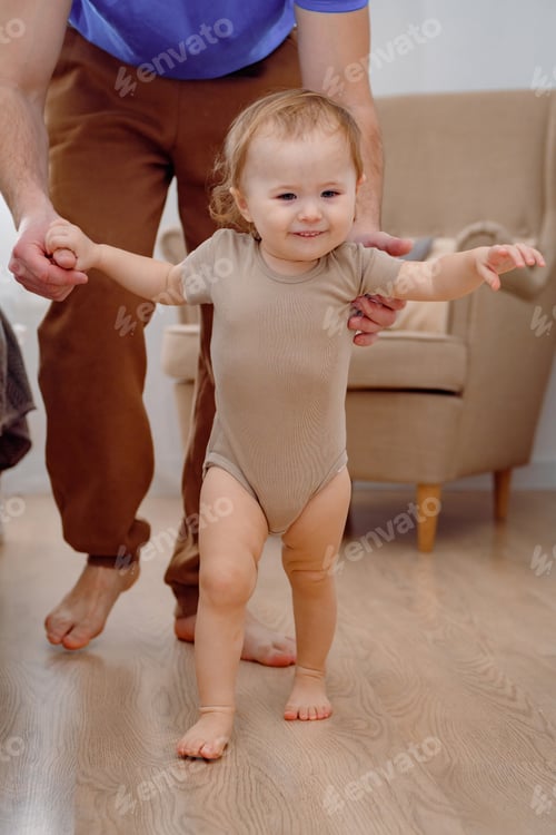 Preview: A cute cheerful smiling baby learning to walk supported by father. First steps of a one year toddler