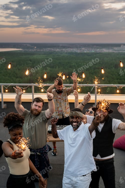 Preview: Joyful young people dancing with bangal lights at rooftop party