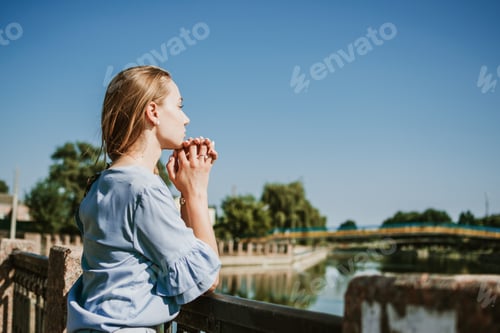 Preview: Lonely pensive Young blonde woman in blue dress standing near the city river in summer sun day