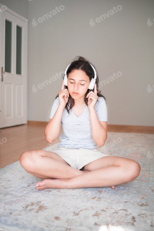 Preview: Pretty young girl listening to music with headphones while sitting on carpet at home