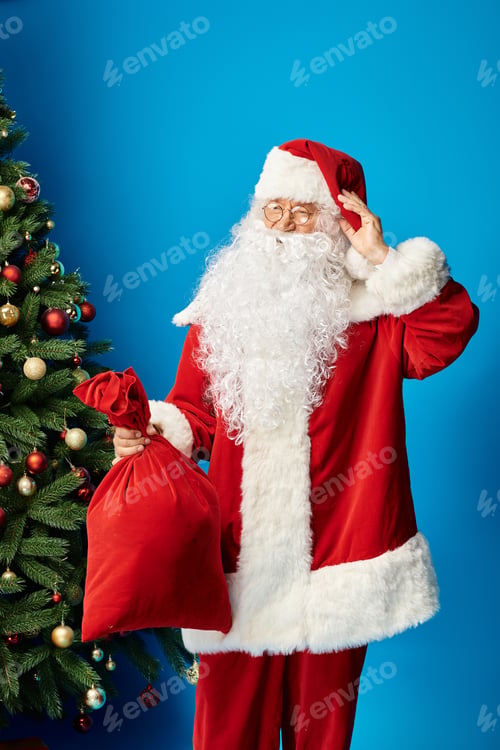 Preview: happy Santa Claus with beard and eyeglasses in red costume holding sack bag with Christmas presents