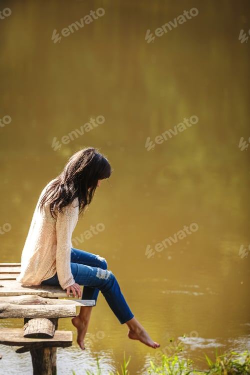 Preview: Young woman sitting on the old bridge barefoot. Summer, green forest, lake and air in the evening