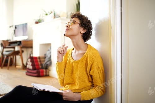 Preview: young african american woman sitting on floor with pen and book thinking