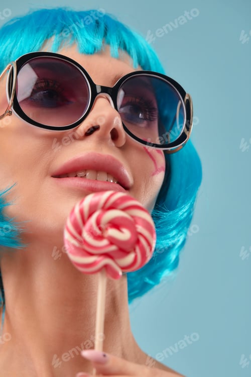 Preview: Young funky girl in blue wig and eyeglasses, holding lollipop candy, looking aside with dreamful