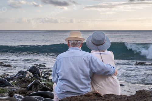 Preview: Rear view of caucasian senior couple sitting on the pebble beach at sunset light admiring horizon