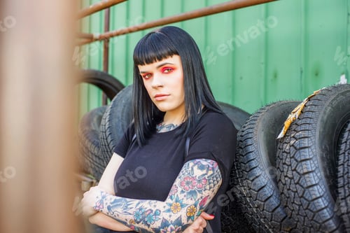 Preview: Tattooed woman leaning against tyres arms crossed looking at camera