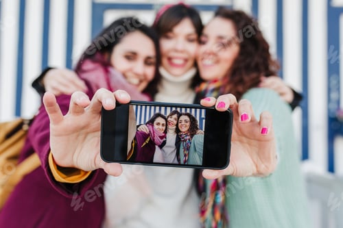 Preview: happy women friends using mobile phone in front of colorful houses.Costa Nova, Aveiro, Portugal