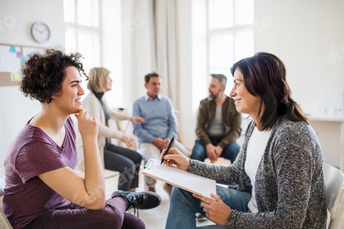 Preview: Senior counsellor with clipboard talking to a woman during group therapy.