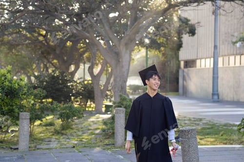 Preview: Adult Asian man wearing black gown cap walking campus holding diploma red ribbon, copy space