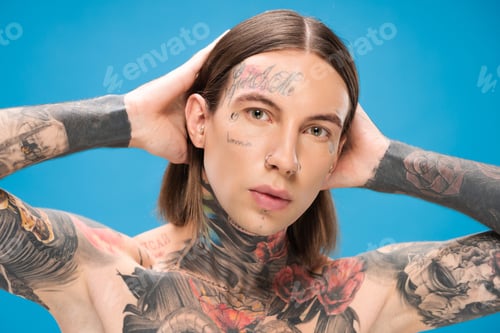Preview: young and pierced man with tattoos looking at camera while posing isolated on blue