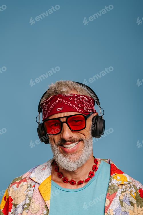 Preview: Joyful senior man in funky shirt and headphones listening to the music against blue background