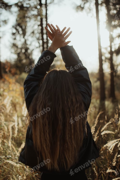 Preview: Vertical shot of a young woman standing in a lush, green forest, her arms raised up towards the sky
