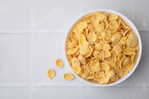 Preview: Breakfast cereal. Tasty crispy corn flakes in bowl on white tiled table, top view. Space for text